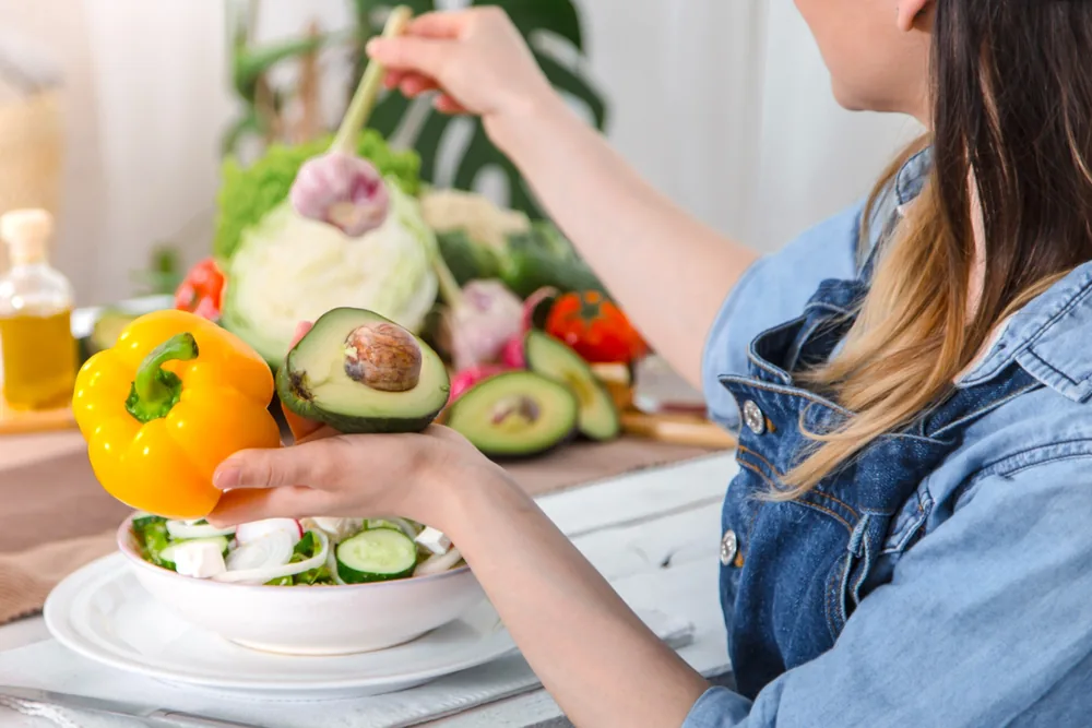 Mujer sentada a la mesa con un pimiento amarillo y medio aguacate frente a un plato de ensalada