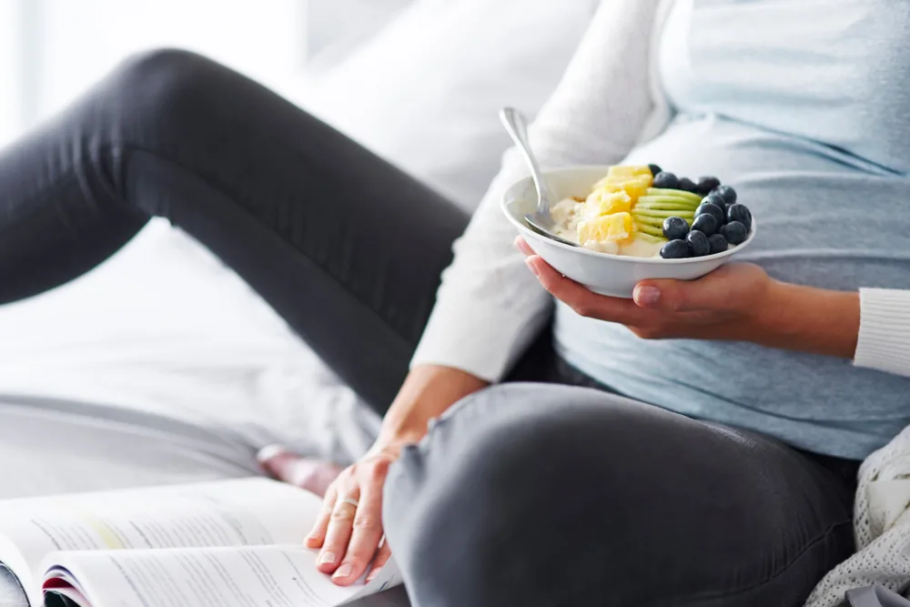 Mujer embarazada sentada con un tazón de fruta y yogur en la mano y una revista abierta a su lado