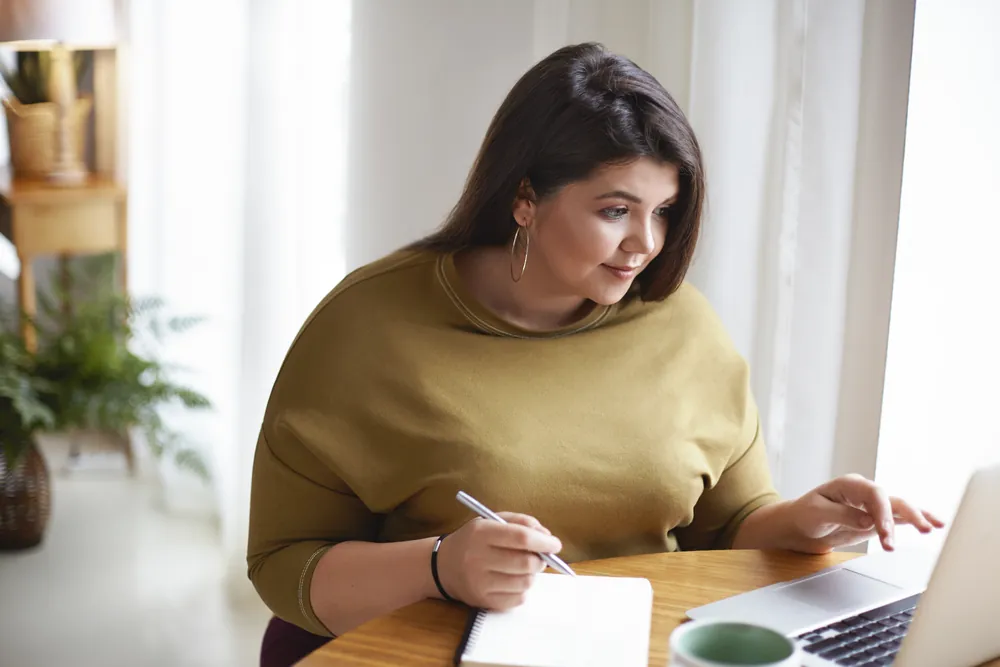 Mujer tomando notas frente a una laptop en una mesa junto a una ventana