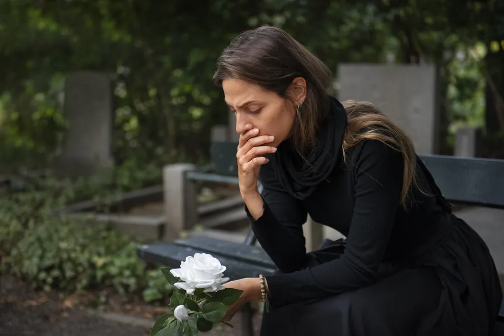 Mujer de luto sentada en una banca de un cementerio con una rosa blanca a su lado